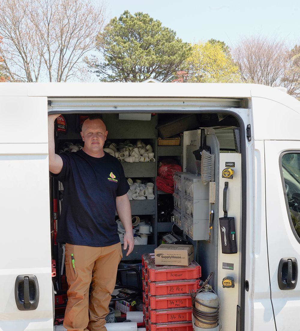 Man stands in open service van, wearing navy shirt and brown pants, tools visible inside.