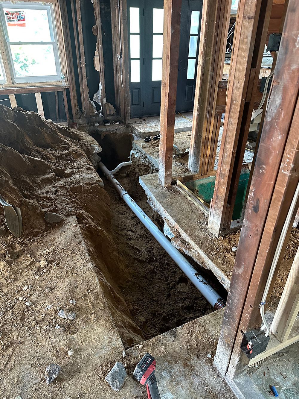 Excavated trench with gray pipe inside, exposed wooden frame of house.