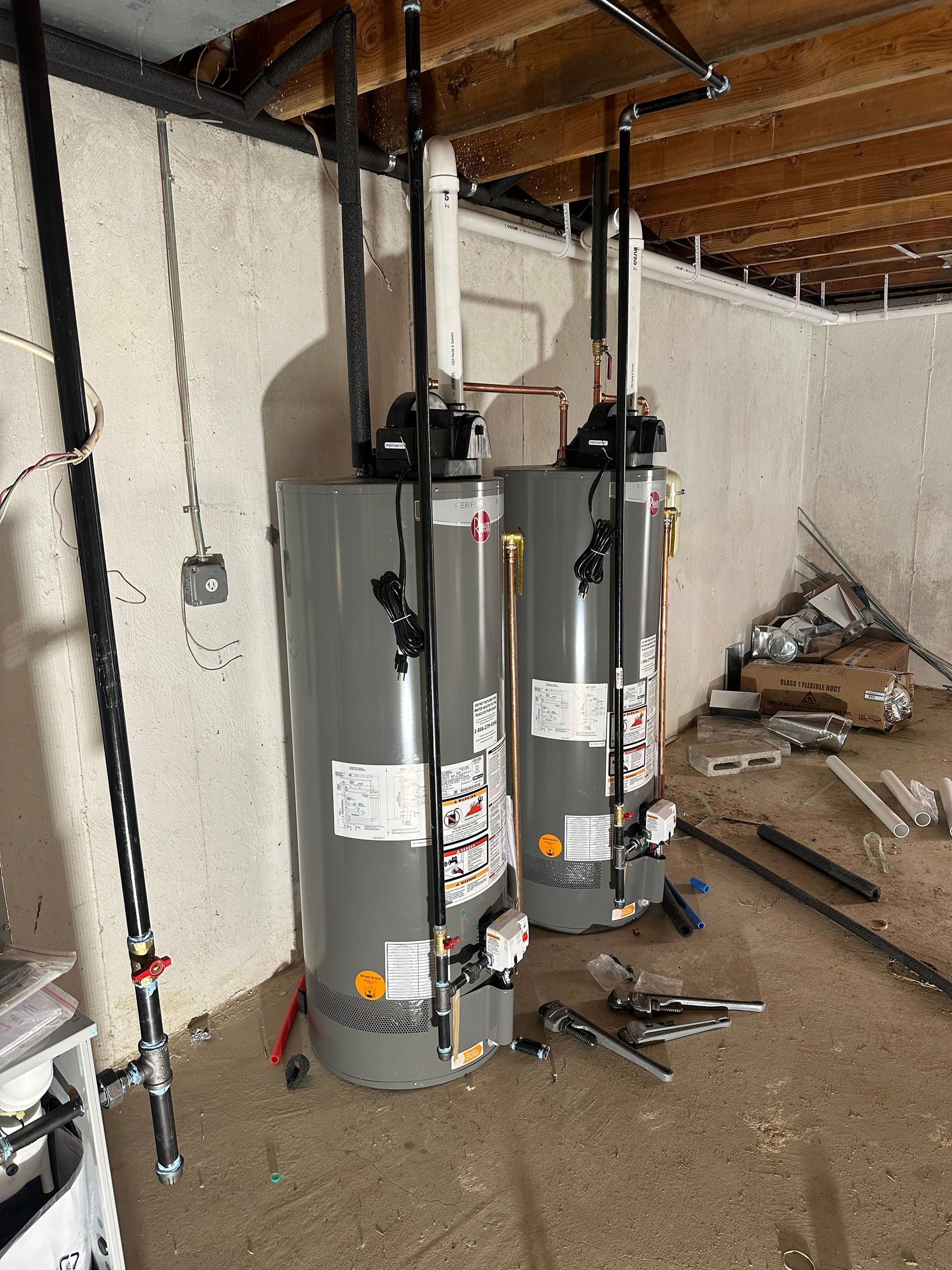 Two tall gray water heaters installed in a basement, with black pipes and a white vent.