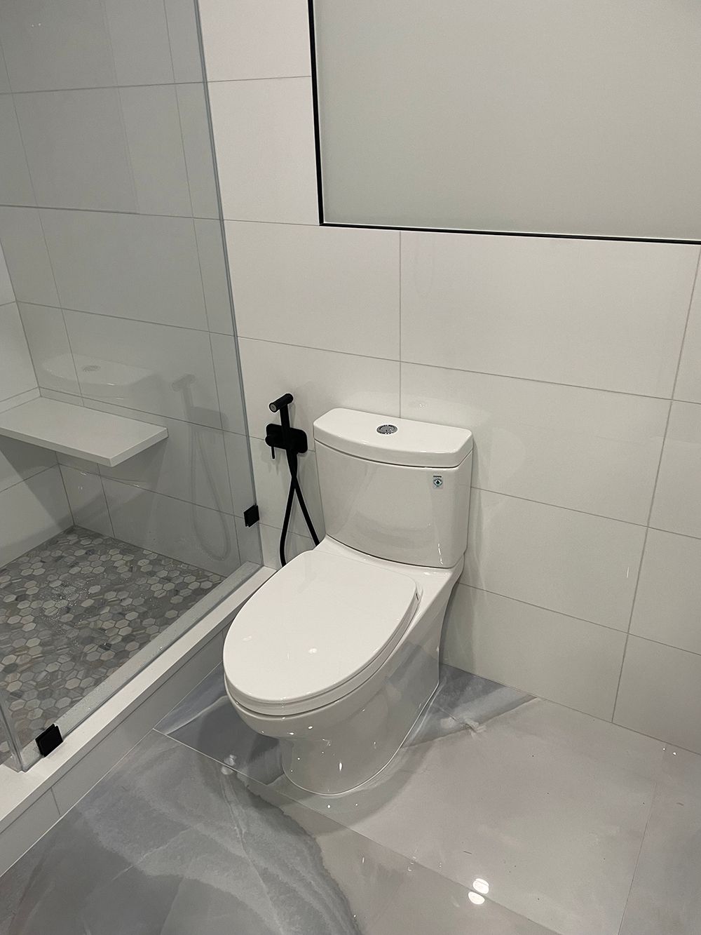 White toilet in a modern bathroom with a glass shower, gray tiled floor, and a black bidet sprayer.