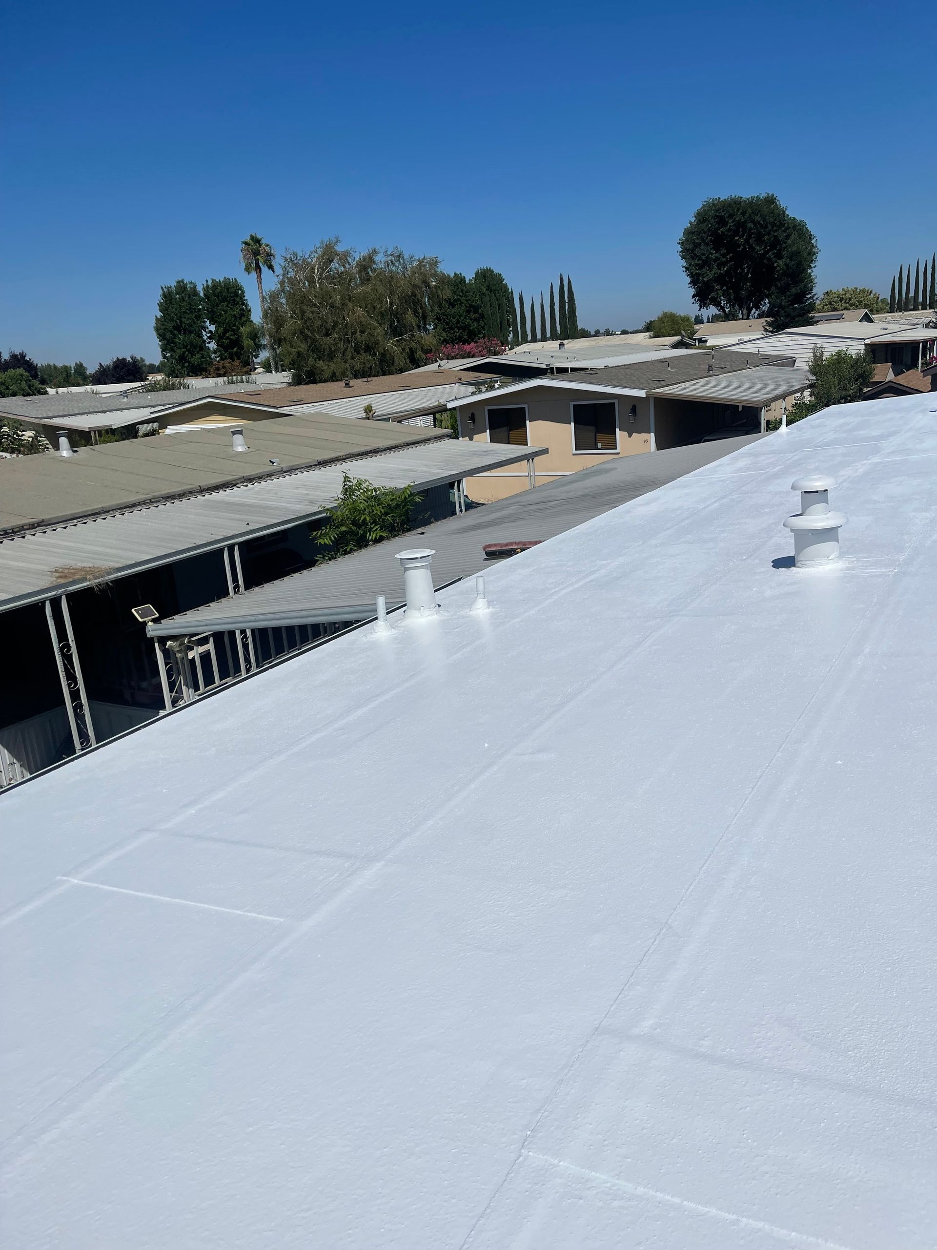 Rooftop with white coating and vents overlooking low buildings and trees under a clear blue sky