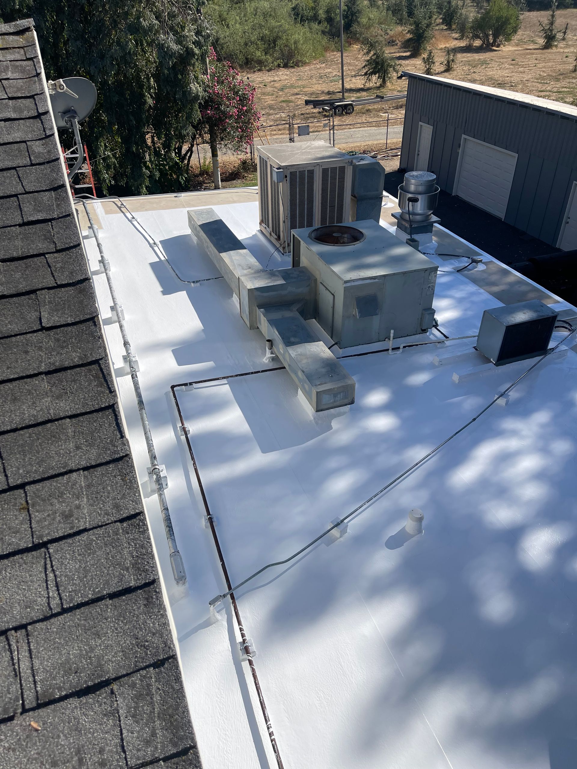 Rooftop HVAC units on a white flat roof beside a dark shingled roof, with trees in the background.