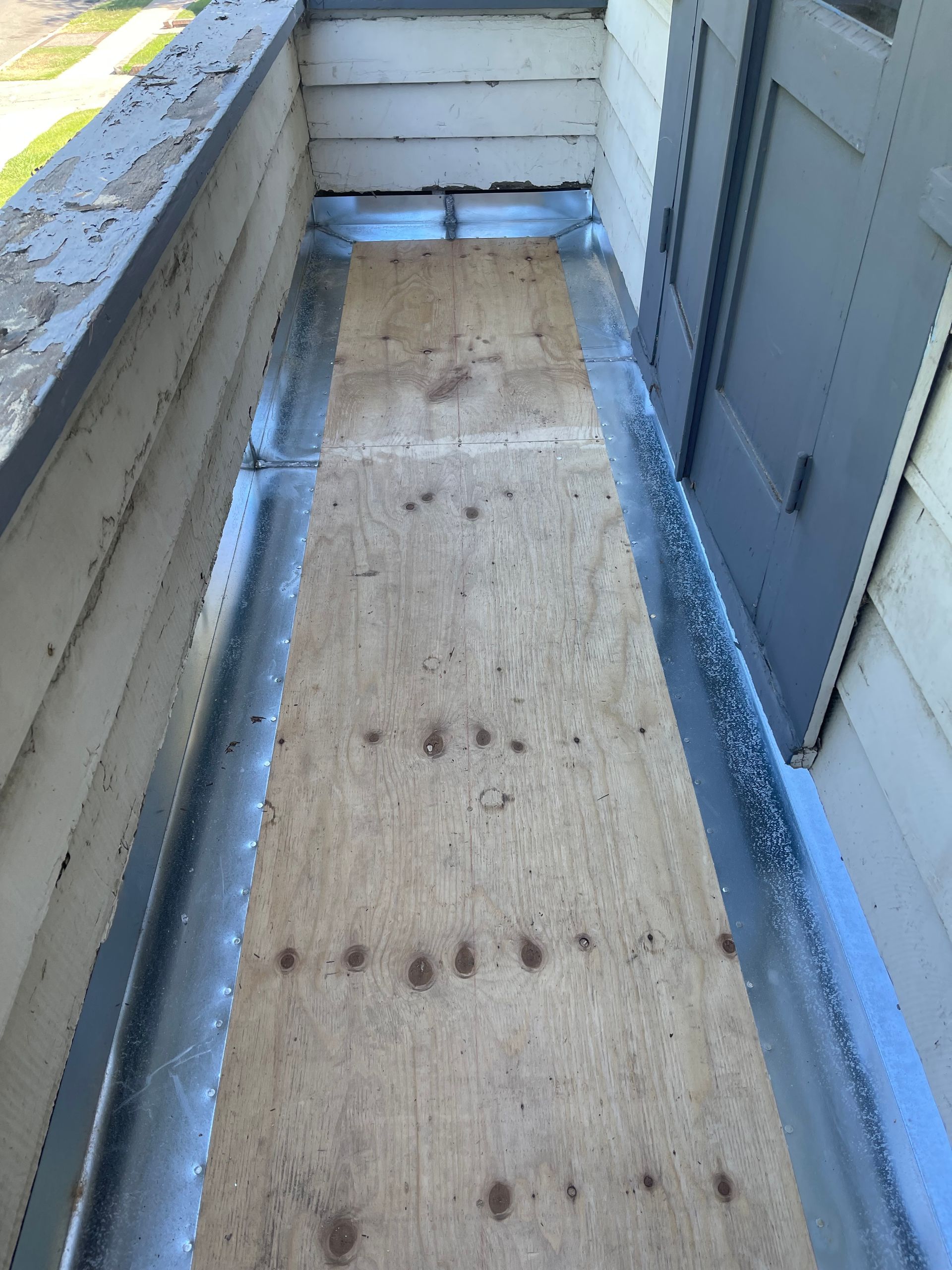 Narrow rooftop walkway with plywood floor, gray walls, and blue edging under daylight
