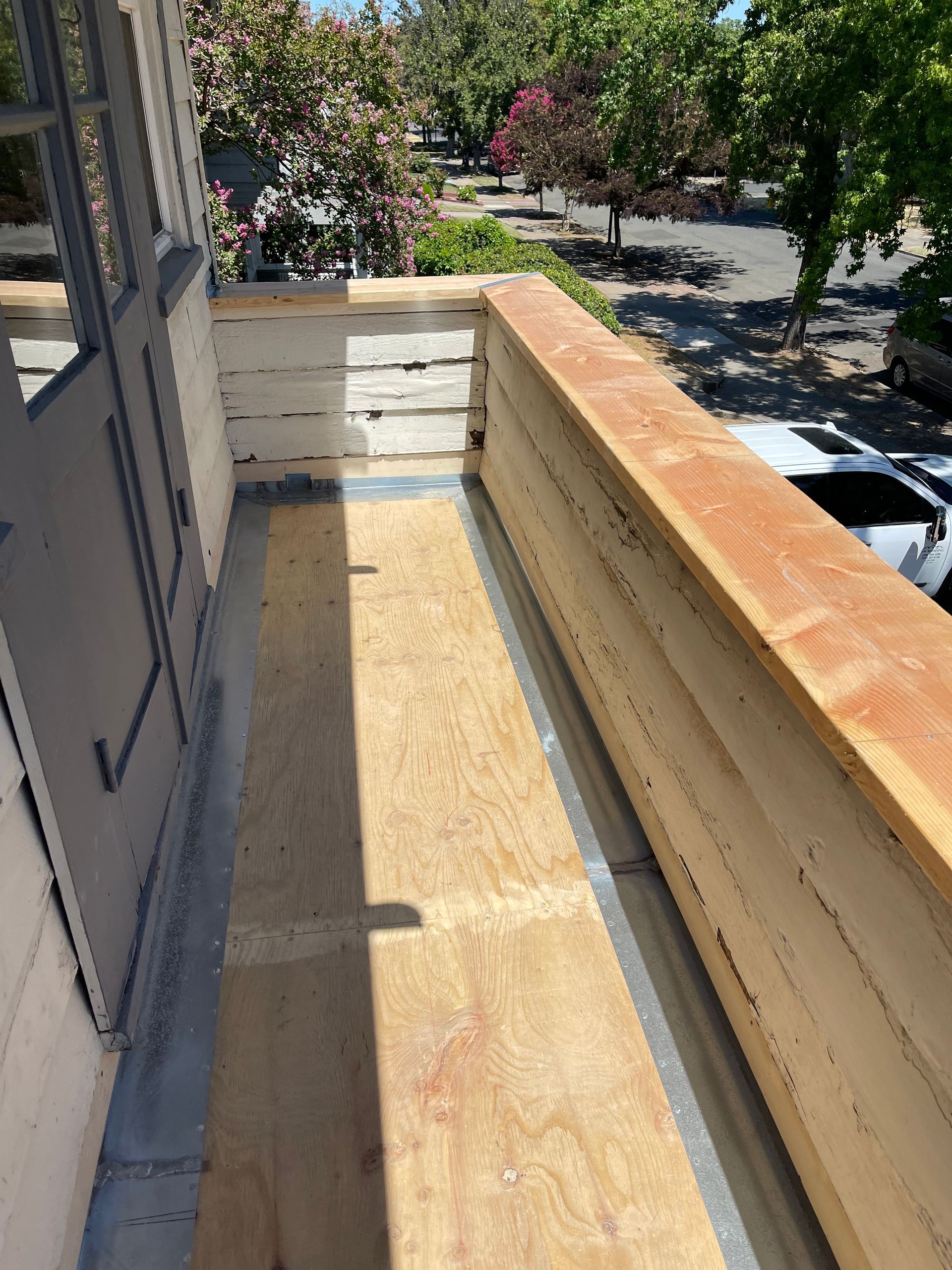 Narrow wooden balcony with unfinished railing and sunlit view of trees and parked cars below