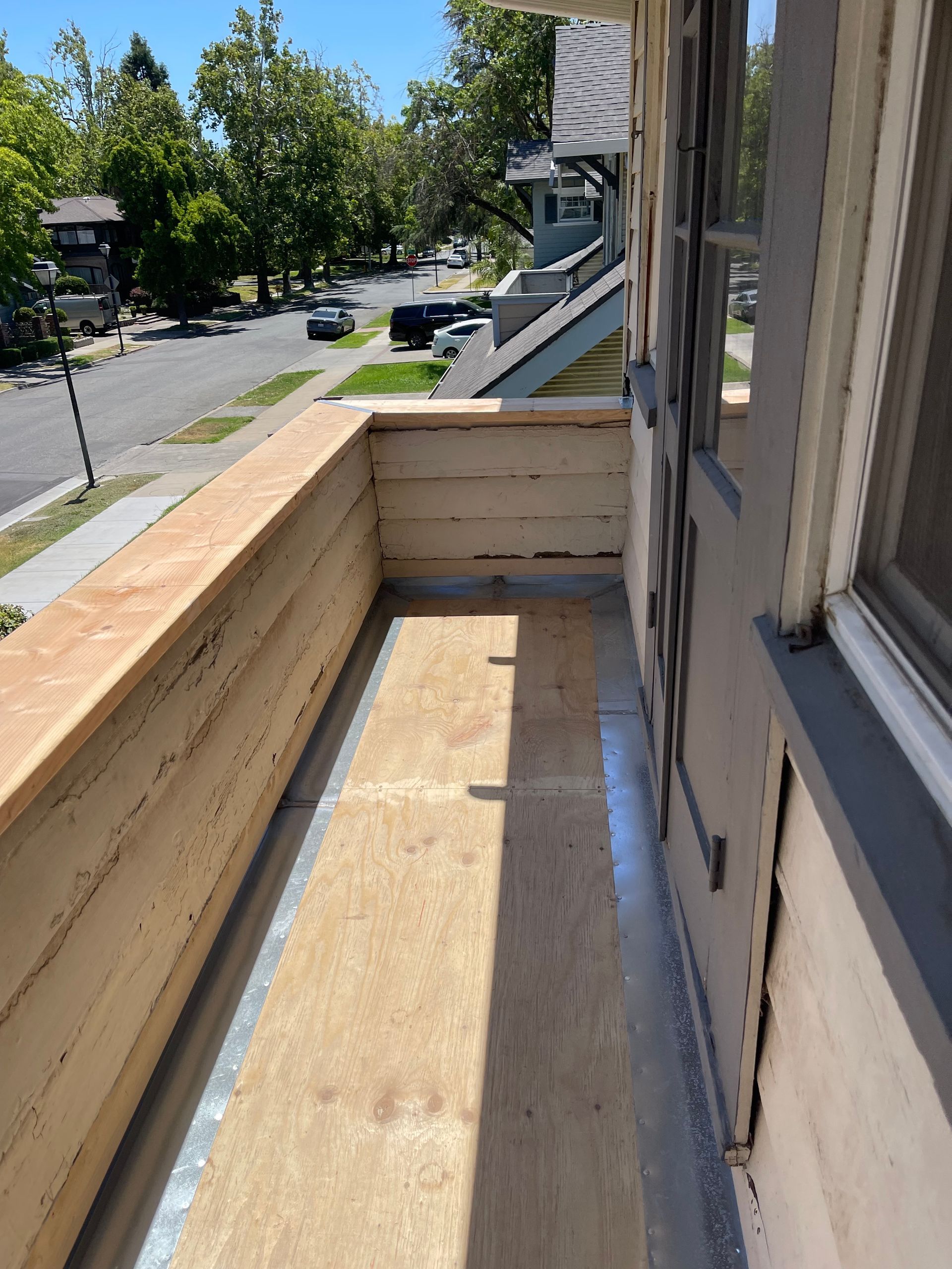 Wooden balcony under construction beside a residential street, with unfinished railing and flooring.