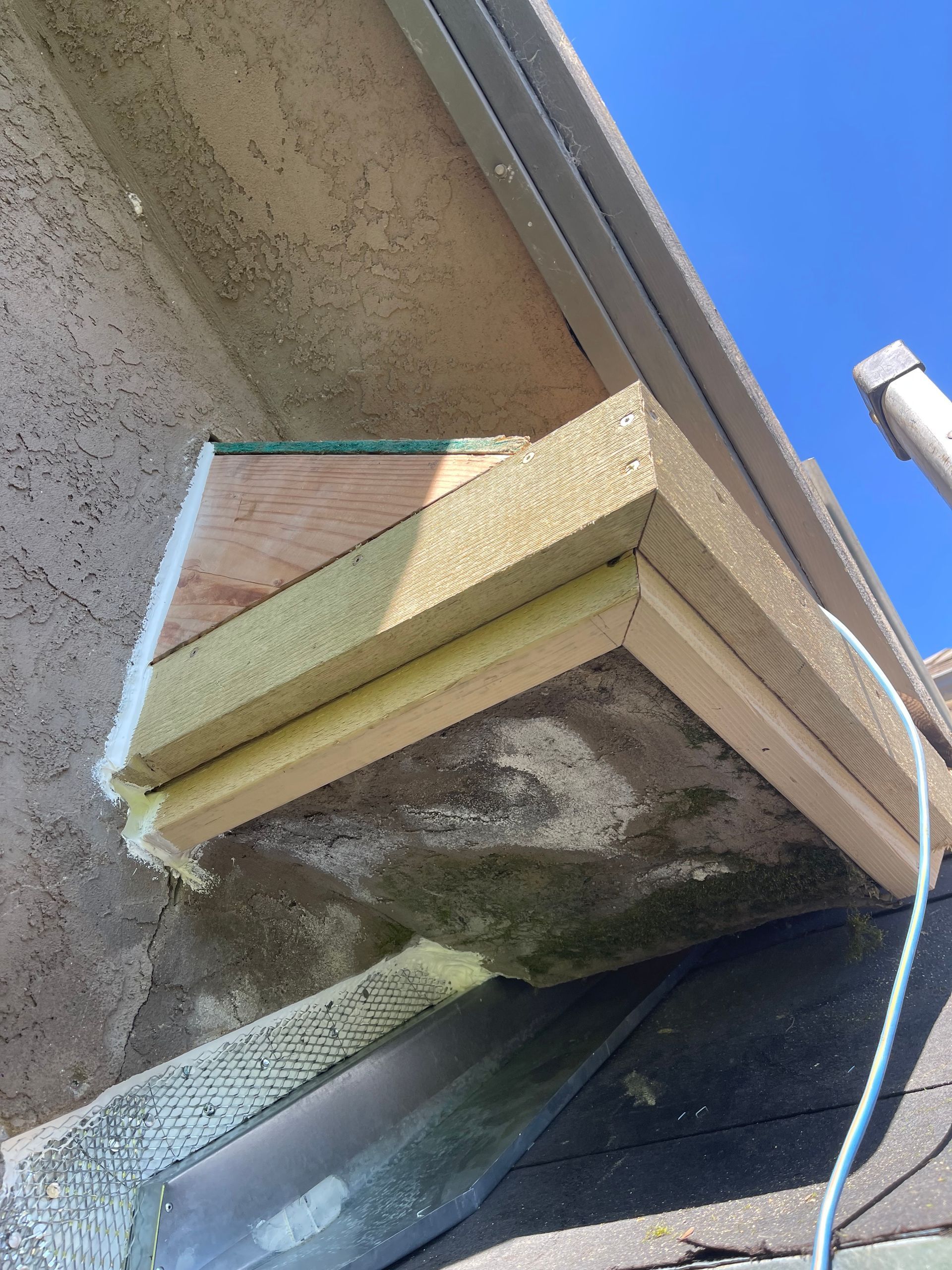 Exterior wall vent hood with blue tape marked around it on a stucco building under a roof edge