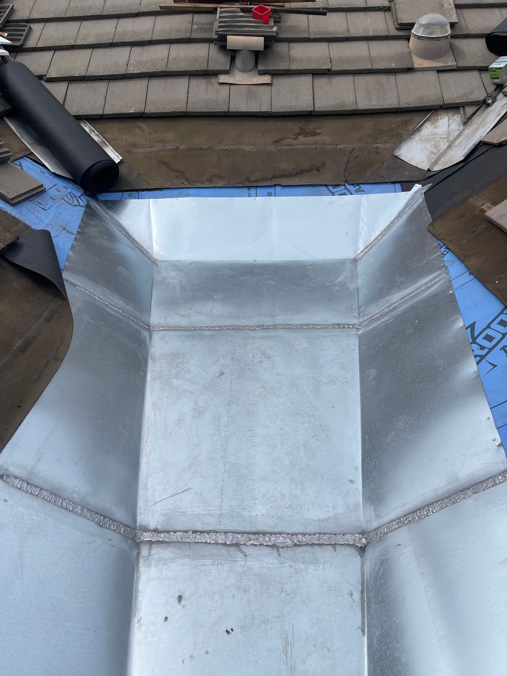Metal roof valley flashing beneath shingles, with blue underlayment and roof vents visible.