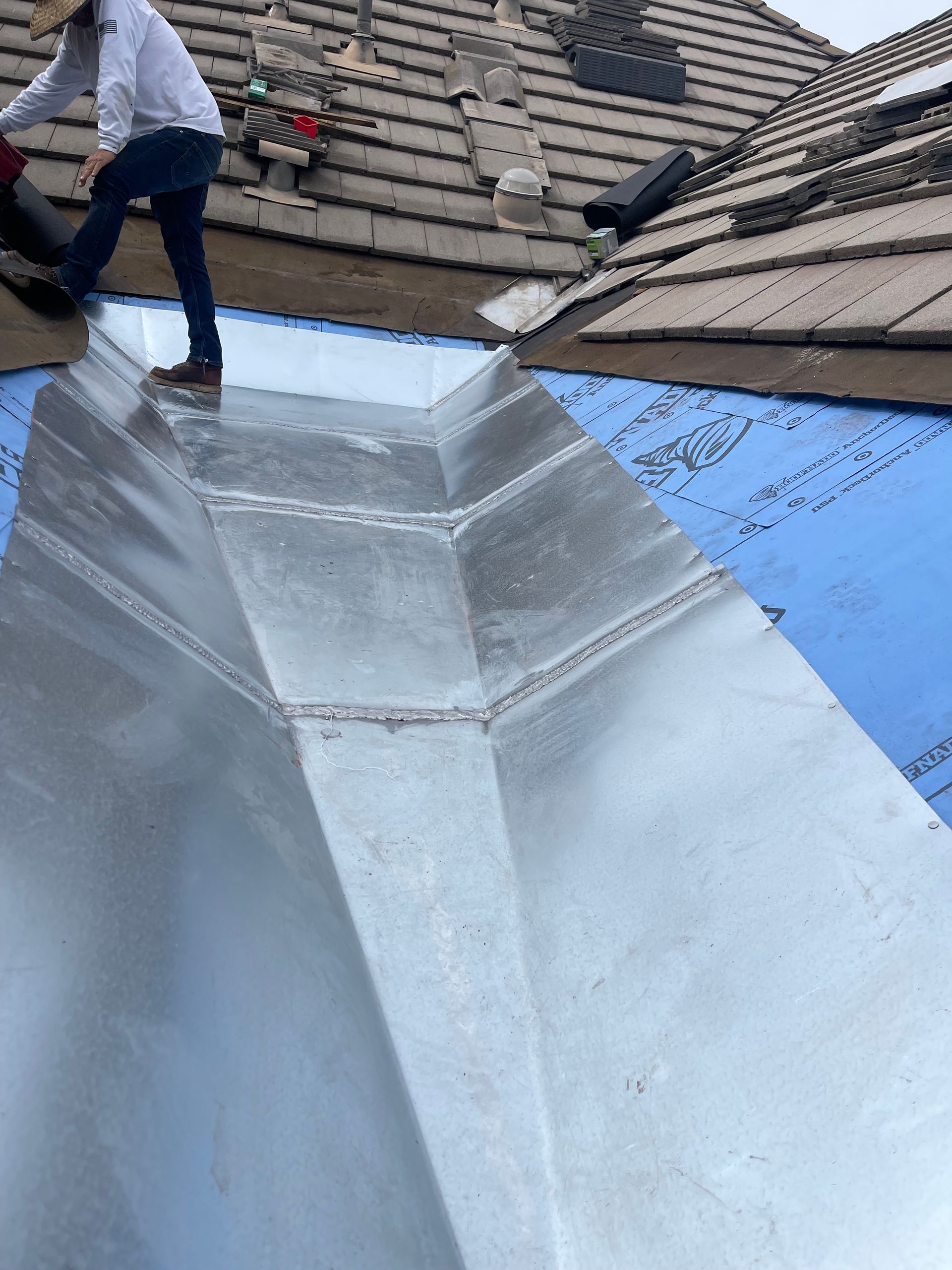Worker on a rooftop inspecting a large metal roof vent surrounded by blue underlayment.