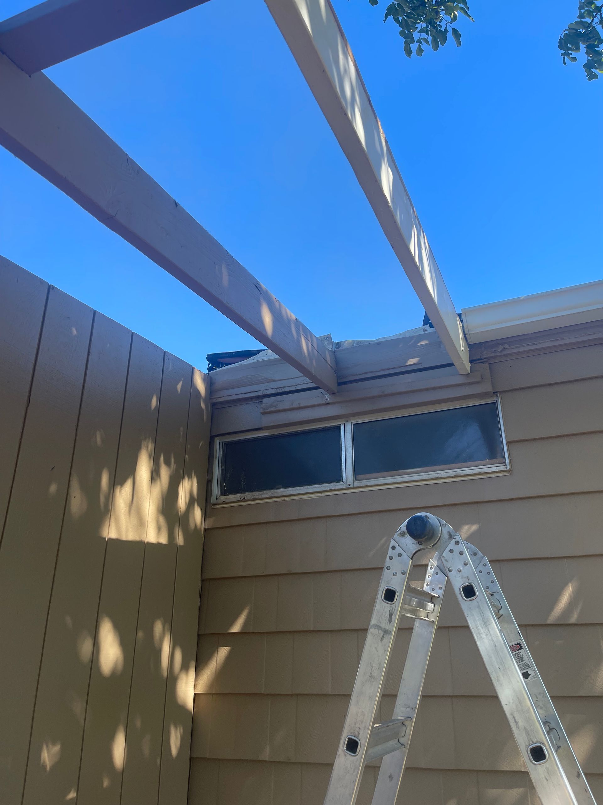 Ladder under white patio beams beside a tan house and window in bright sunlight