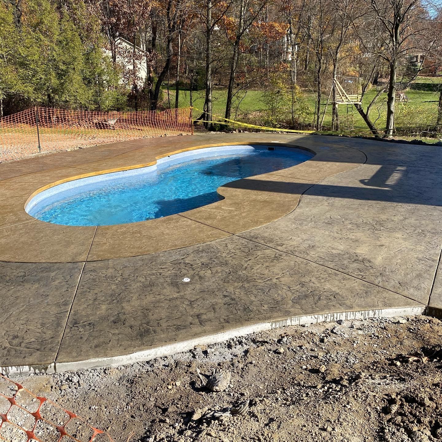 Kidney-shaped backyard pool with bright blue water on a concrete patio, surrounded by trees and grass.