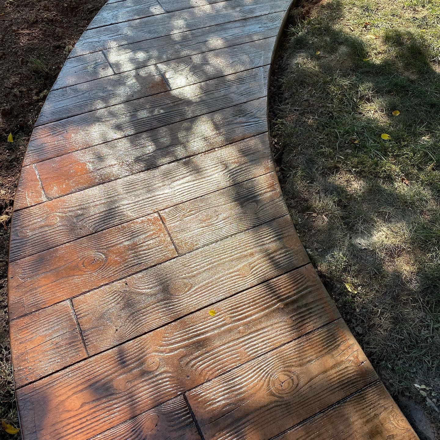 Curved wooden boardwalk with sunlit planks beside grass and dirt