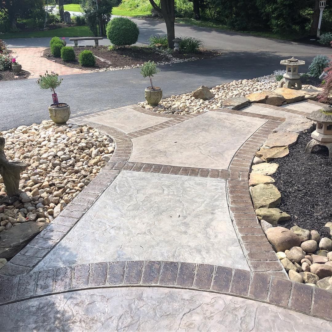 Curved concrete garden path with brick accents, lined by rocks, leading to a driveway and shrubs.