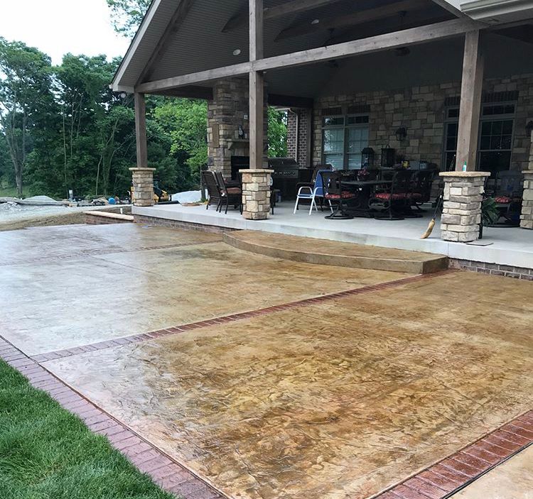 Covered stone patio beside a pavilion with columns and outdoor seating, next to a wet concrete driveway