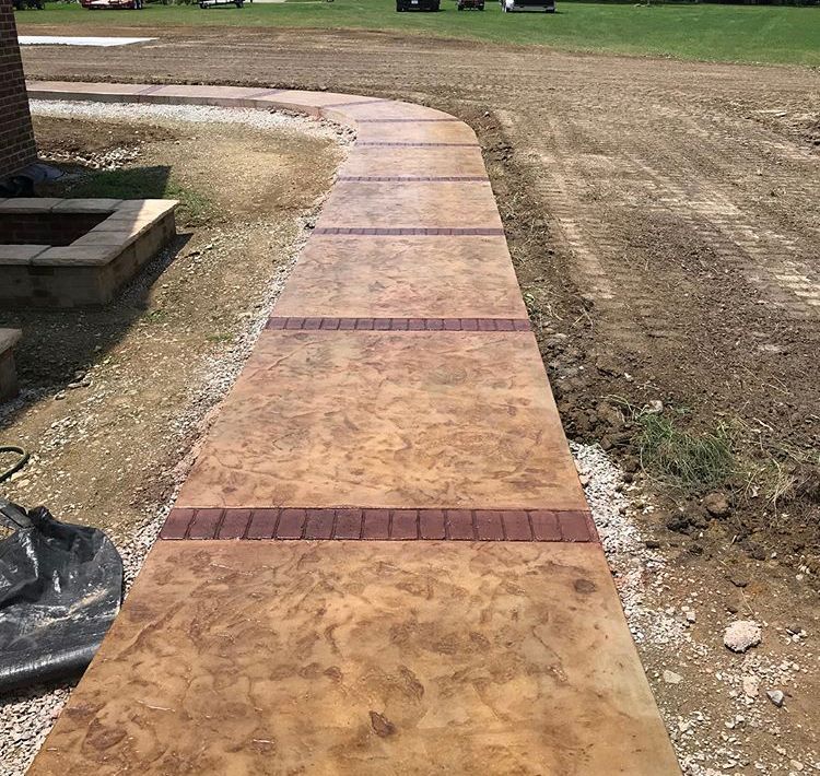 Curved stamped concrete walkway with brick accents beside a grassy yard and patio