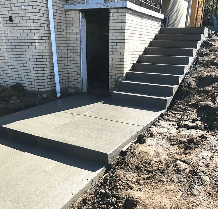 Concrete walkway and steps beside a brick building, with a dirt slope on the right.