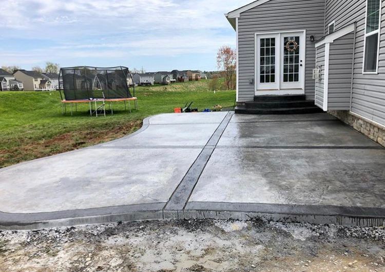 Concrete patio and walkway beside a gray house, with a trampoline on the lawn in the background.