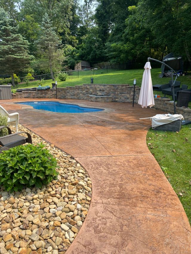 Backyard patio with a small blue pool, stone edging, and a closed white umbrella amidst greenery