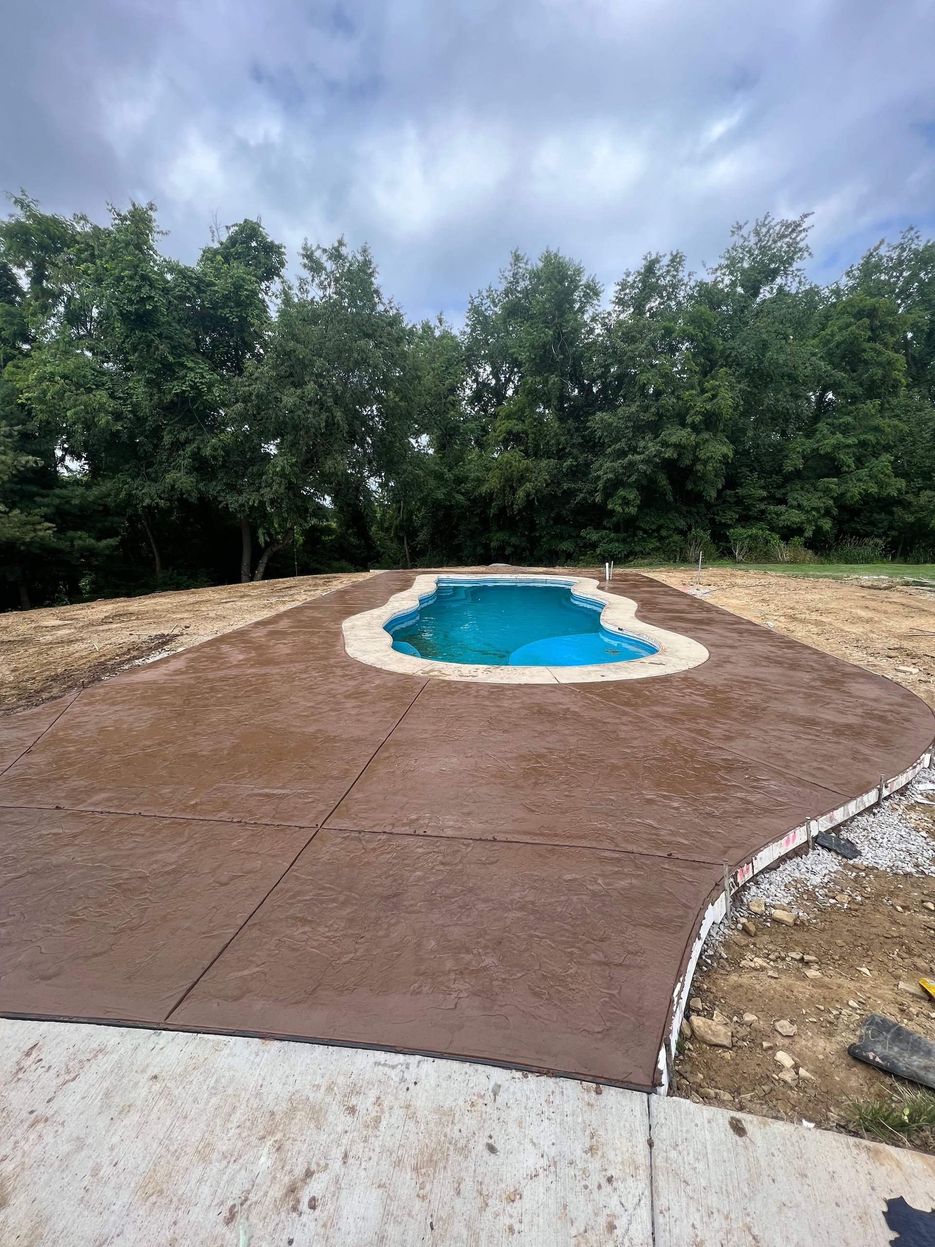Small backyard pool surrounded by a brown concrete deck and trees under a cloudy sky