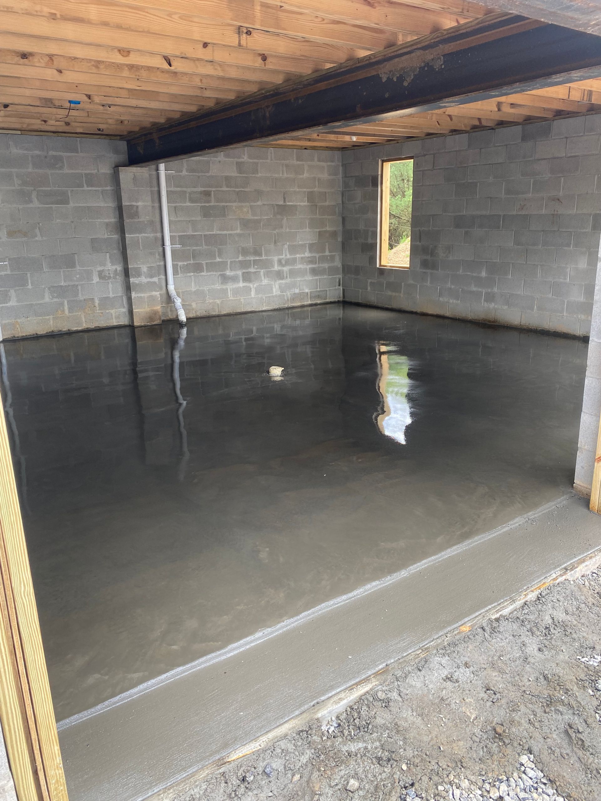 Large empty cinder block room with a freshly poured concrete floor and exposed wooden ceiling beams.