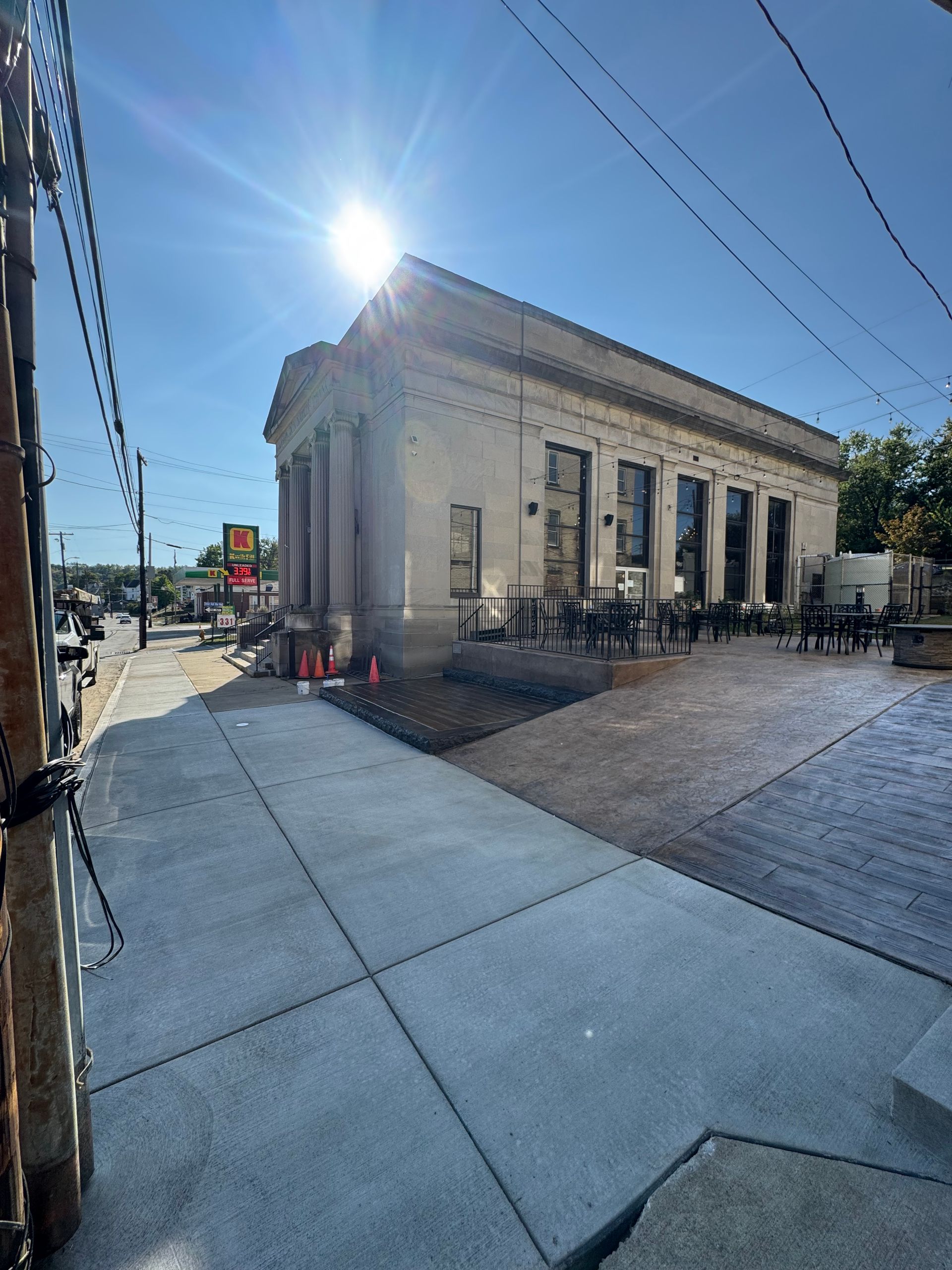 Sunny street corner with a stone building, sidewalk seating, and parked cars under a clear blue sky
