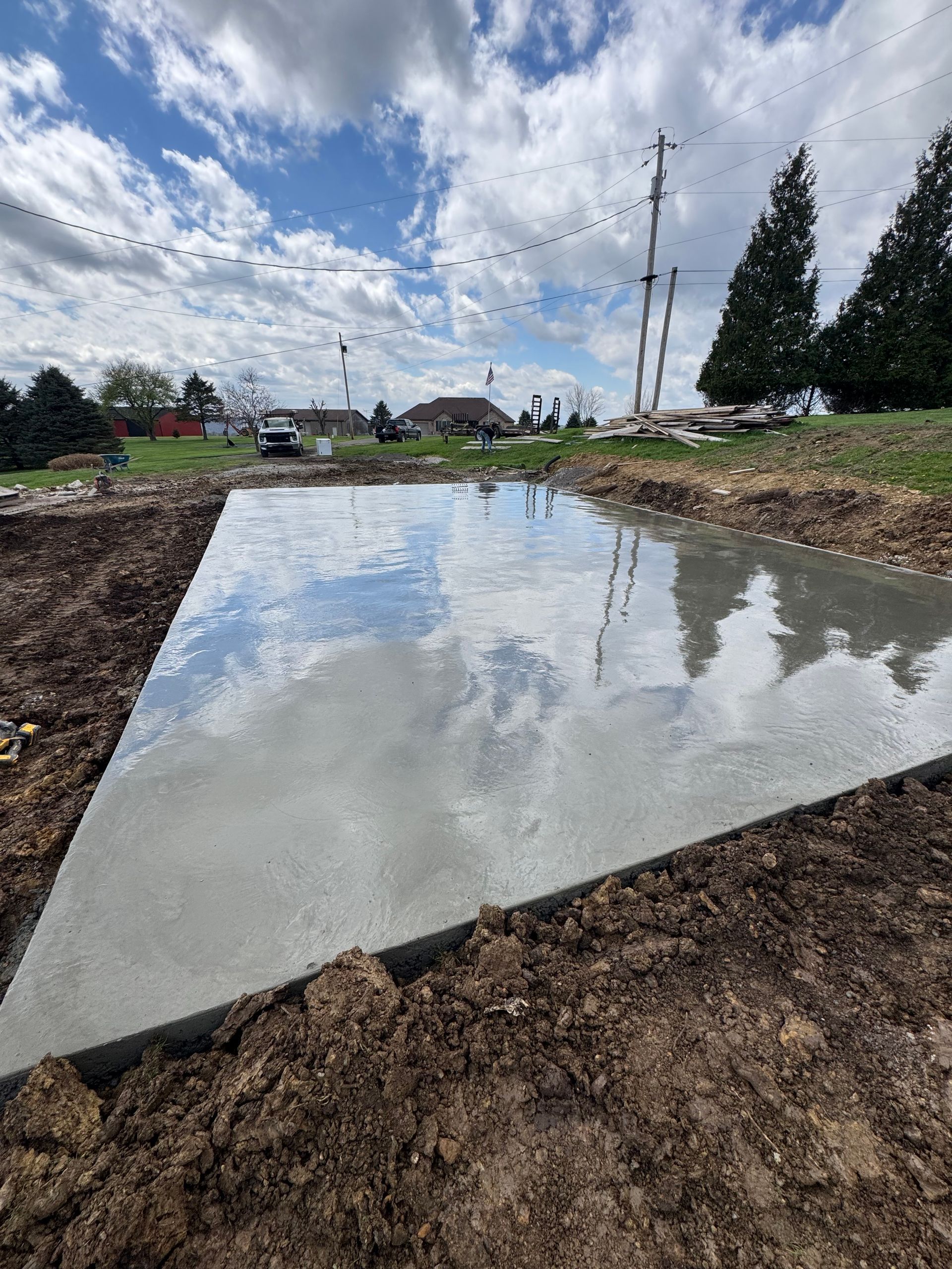 Concrete foundation in a dirt lot reflecting cloudy sky, with utility poles and trees in the background
