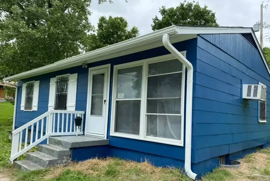 Blue house with white trim, front porch, and white railing in a grassy yard under trees