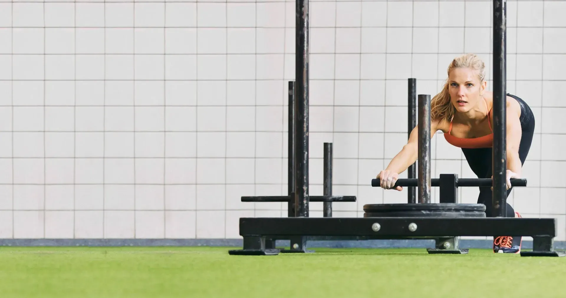 Woman pushing a weight sled across a green floor in a gym.