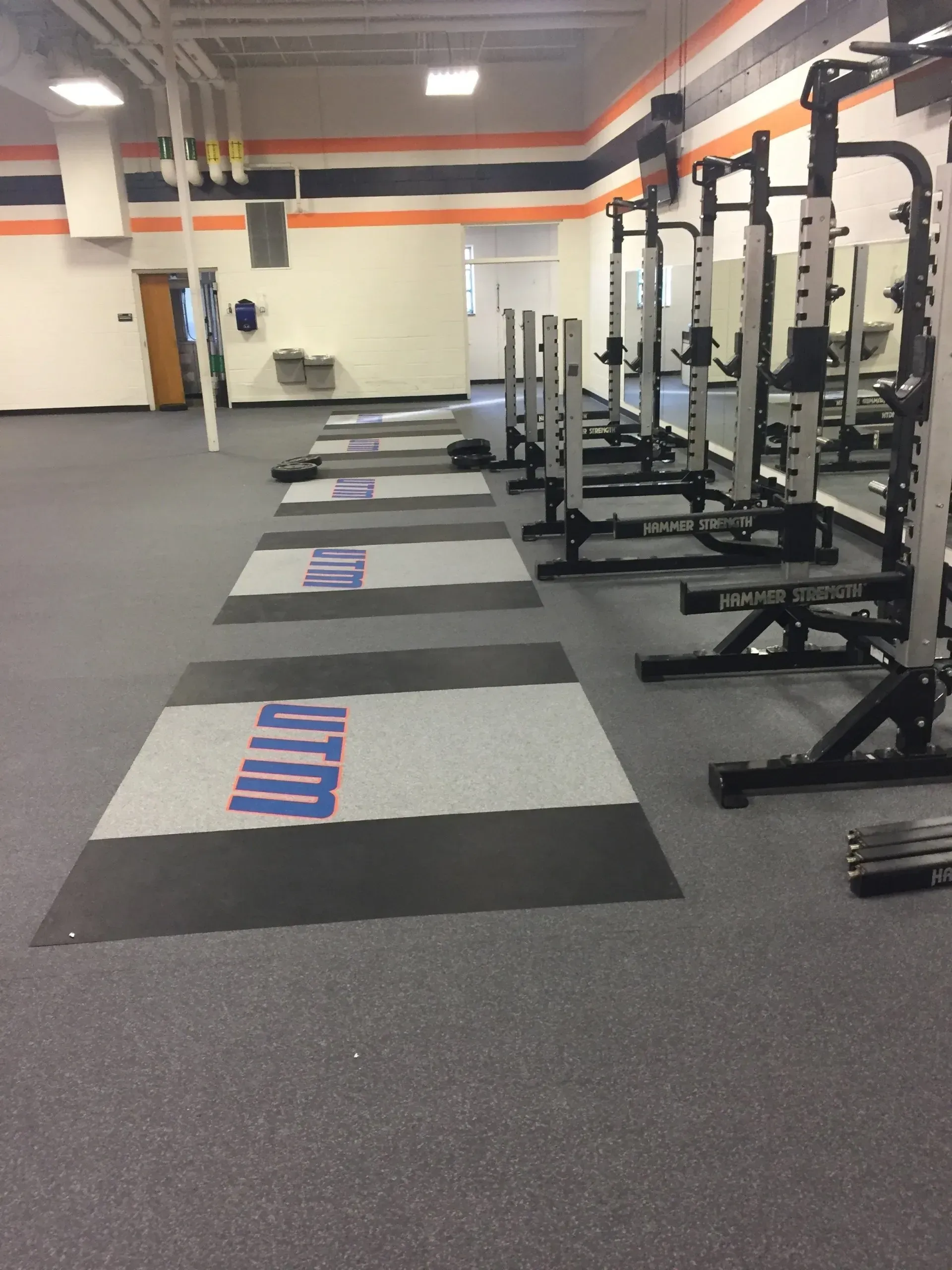 Weightlifting gym with squat racks and logo-covered mats. Orange, blue, and gray color scheme.