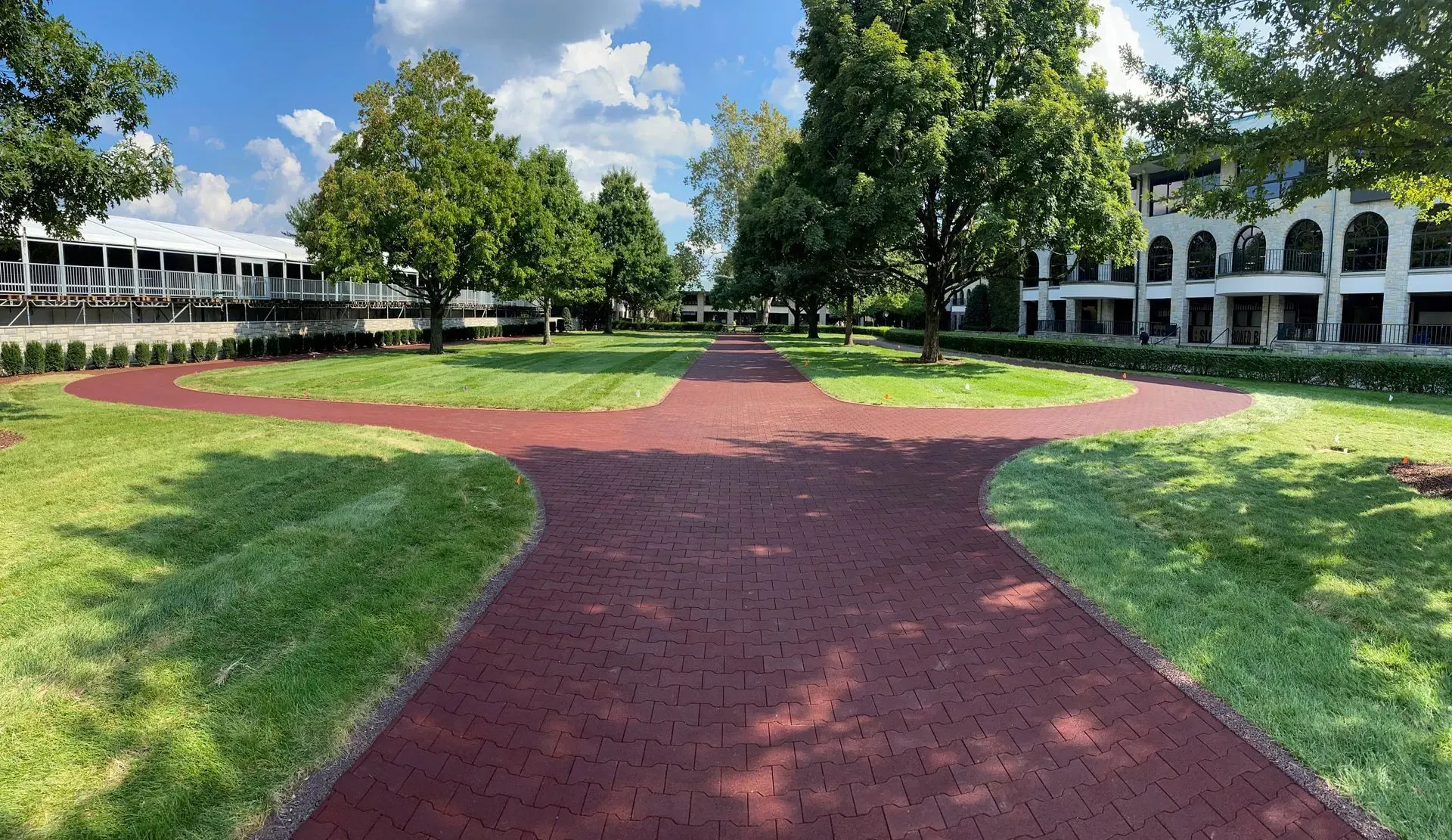 Red brick pathways through a grassy park area with trees and white buildings under a blue sky.