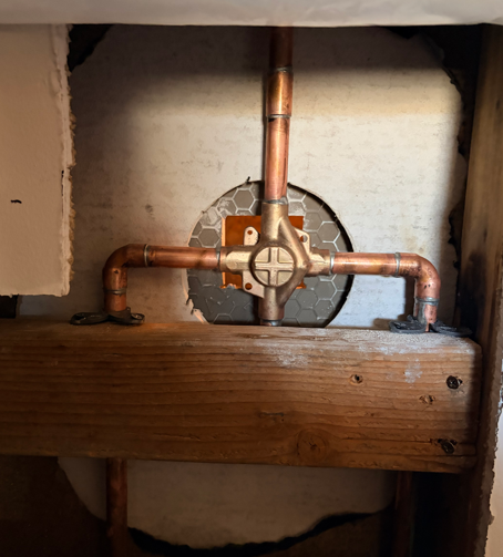 Copper plumbing pipes and a valve inside a wall opening behind a wooden beam.