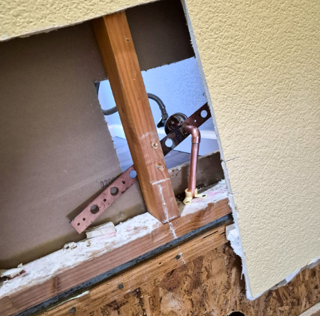 Damaged wall opening exposing wooden framing, pipes, and plumbing inside a beige exterior wall.
