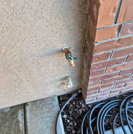 Outdoor wall with two water spigots beside a brick corner and coiled black hose on gravel.