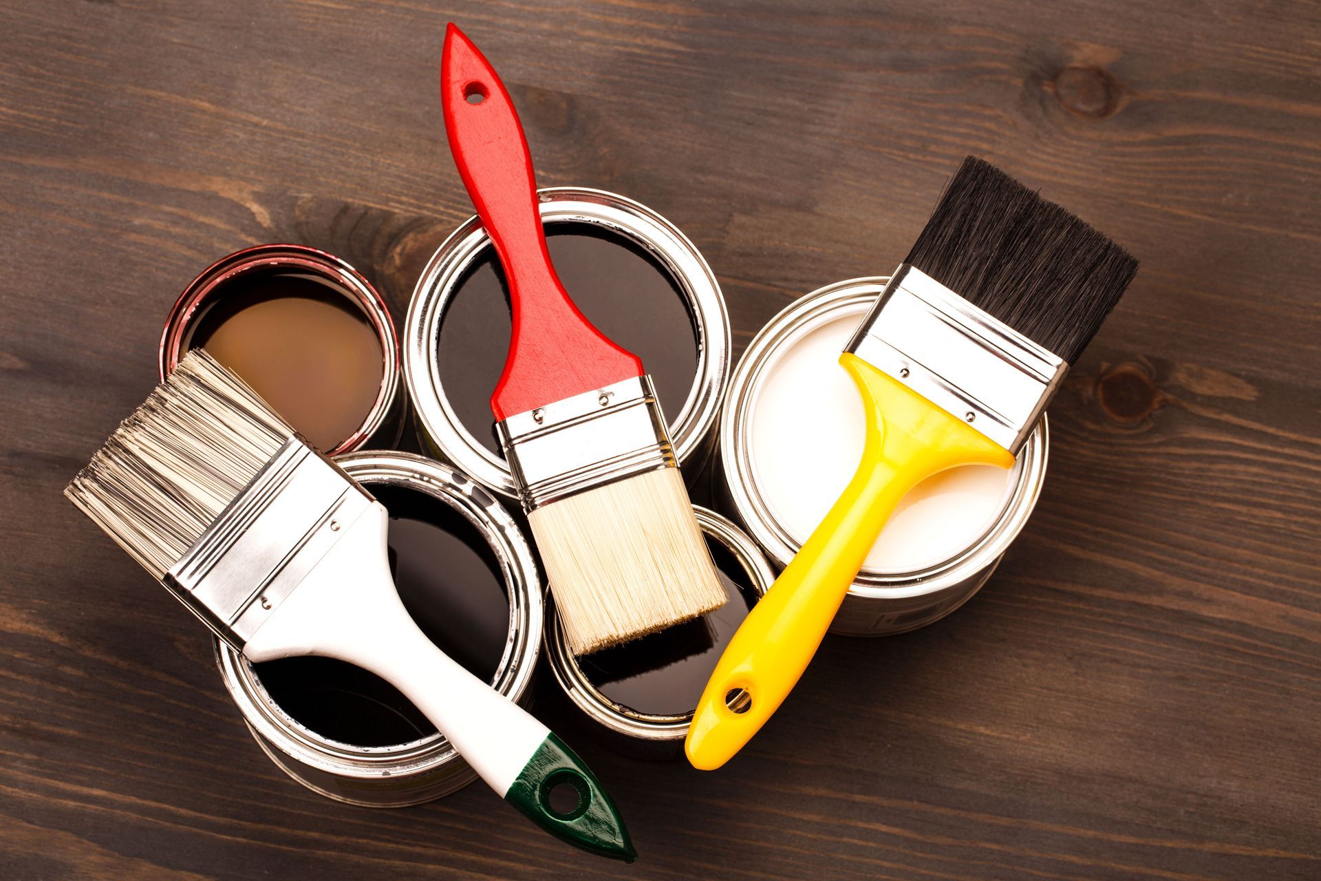 Paintbrushes rest on open paint cans with various dark and light colors on a wooden surface.