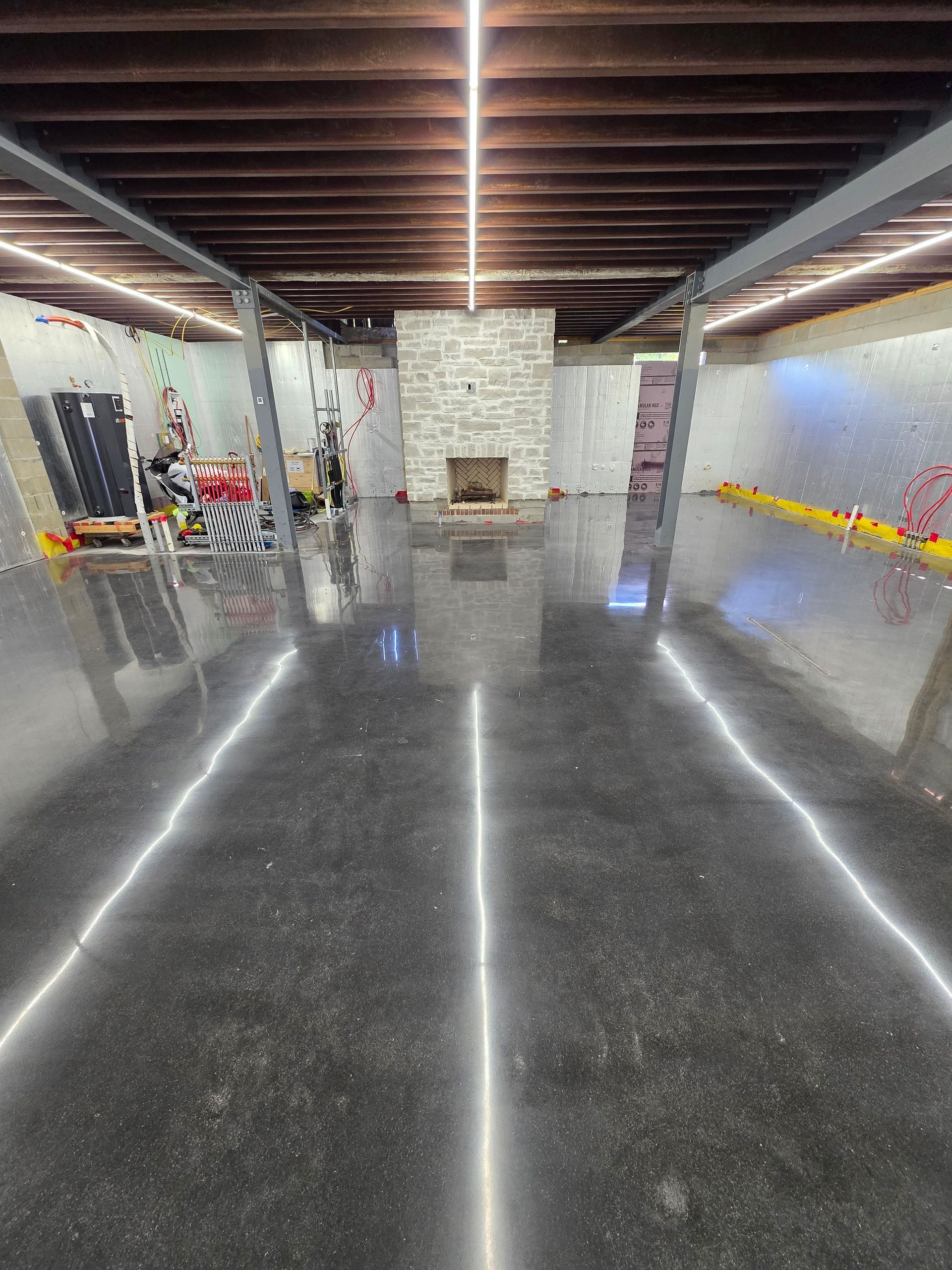 Spacious basement room with polished concrete floor, exposed beams, and a stone fireplace at the far wall