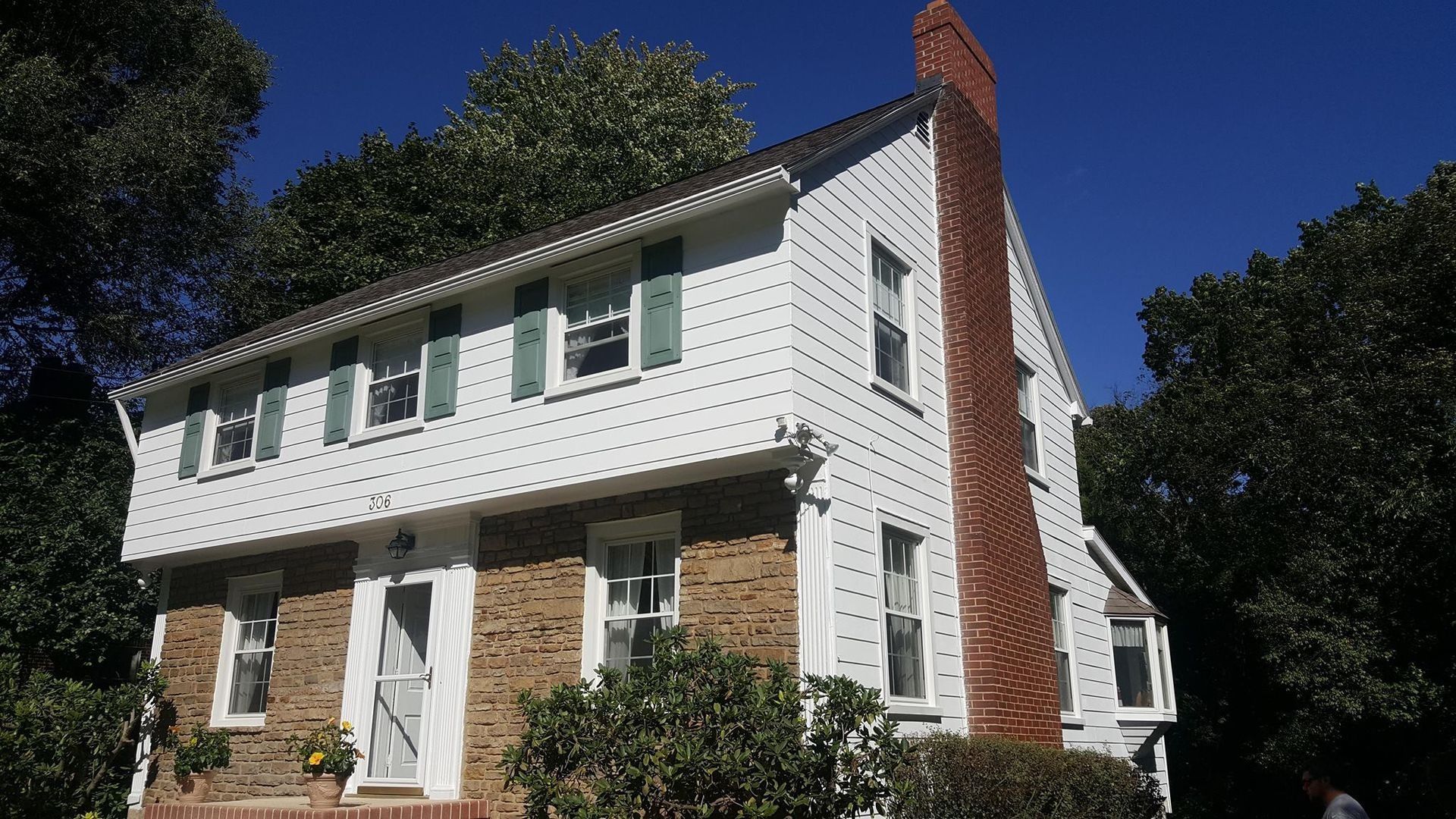 Two-story white house with green shutters, brown brick chimney, and stone facade, under a clear blue sky.