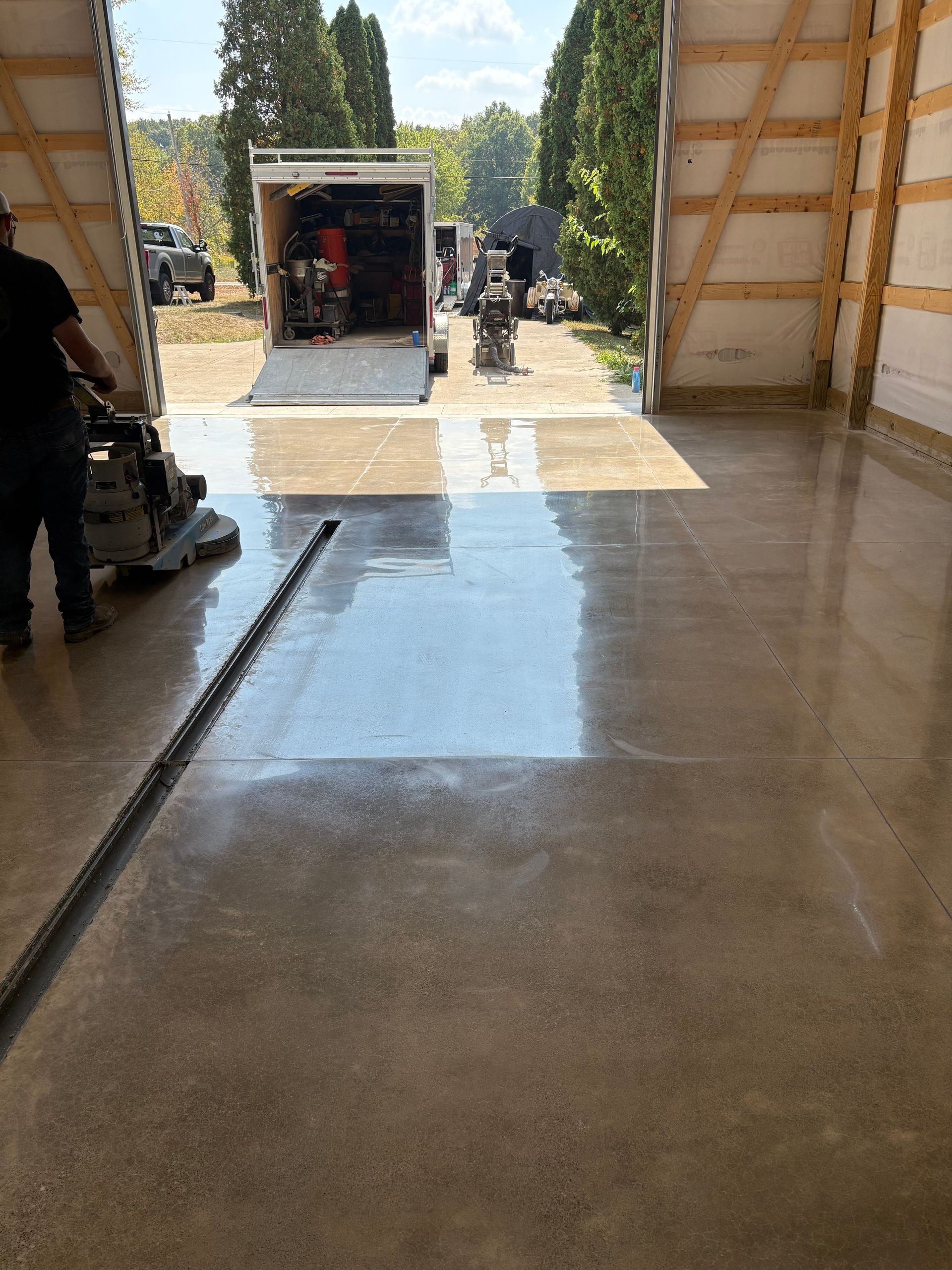 Man grinding concrete floor inside a large building with open doors, trailer parked outside.