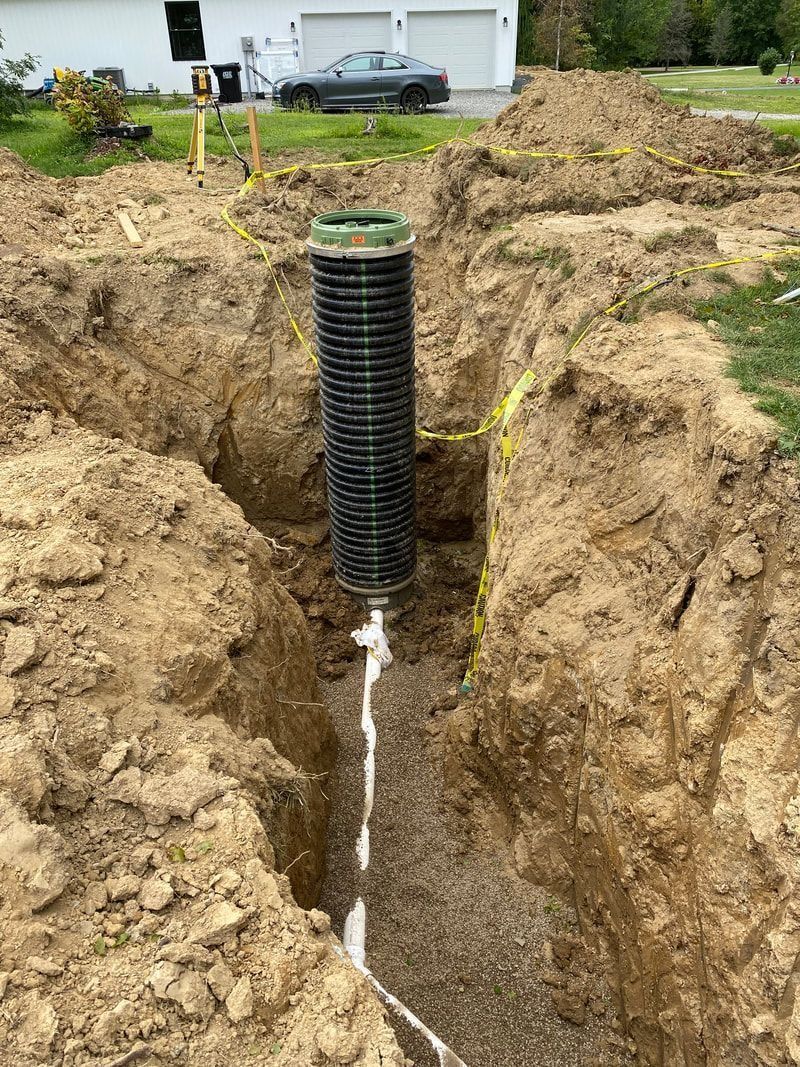 Black drainage pipe in a trench; white pipe extends from it. Dirt surrounds the trench in a yard setting.