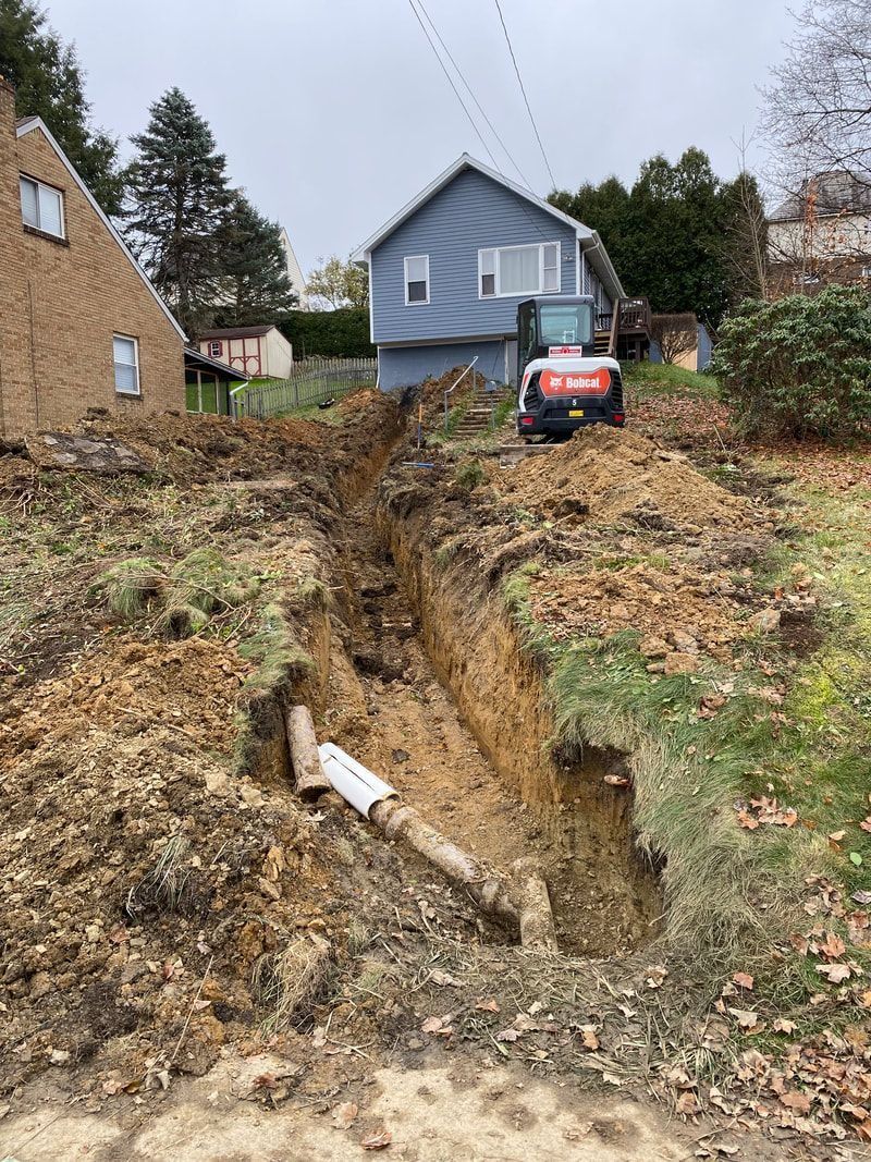 Trench dug near houses, exposing a pipe. A small excavator sits at the end. Overcast day.
