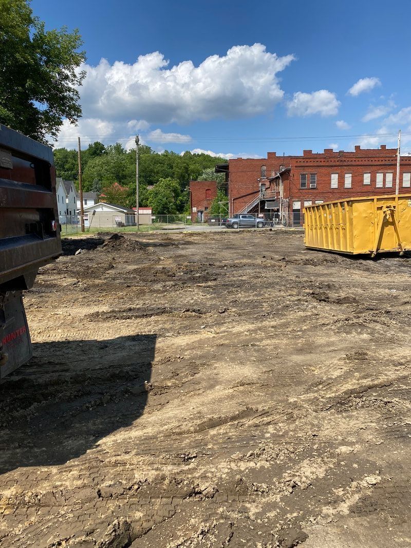 An excavated lot with a yellow dumpster, with a red brick building in the background under a blue sky.