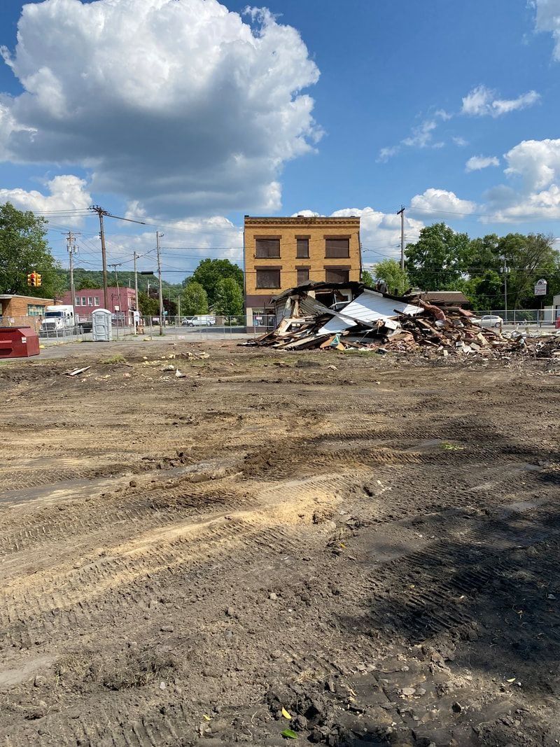 Demolished building site with debris pile. Yellow brick building in background. Blue sky, white clouds.