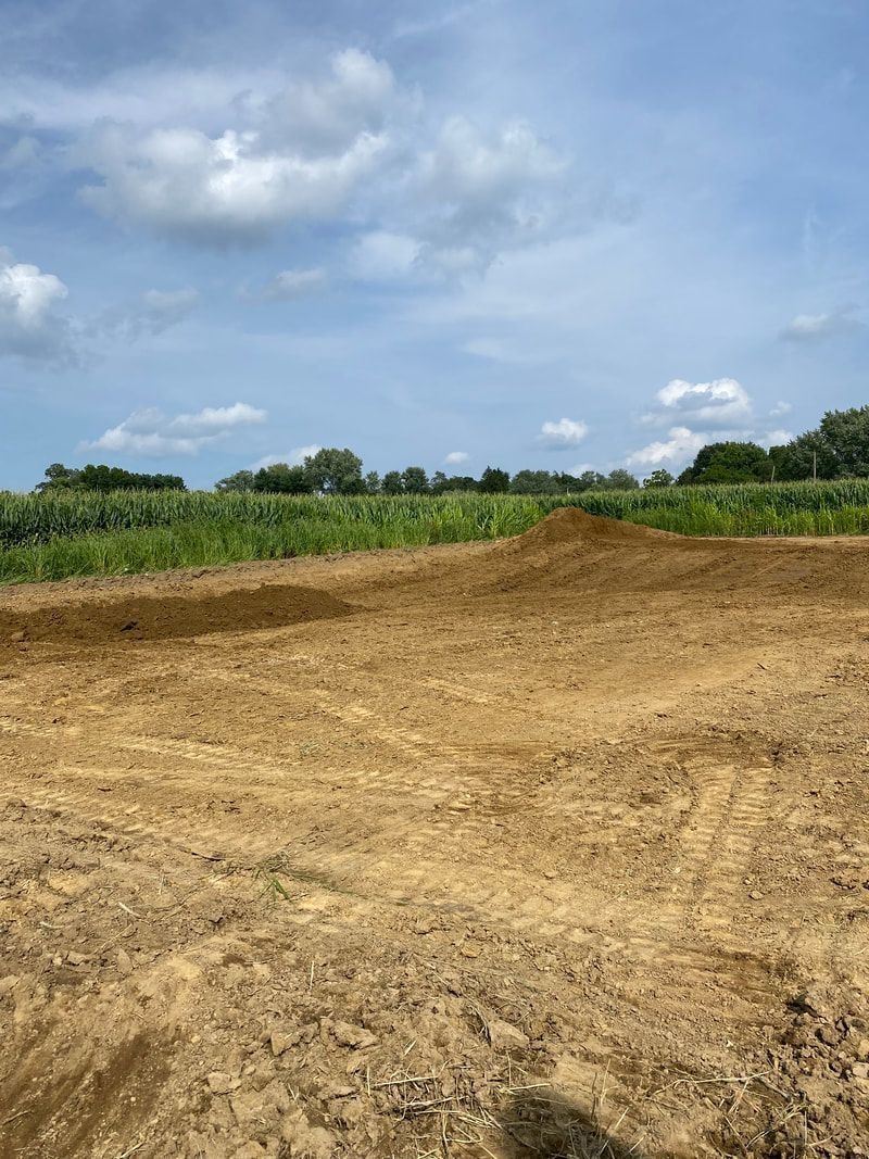 Brown dirt field with a small mound, green crops in the background, under a partly cloudy blue sky.