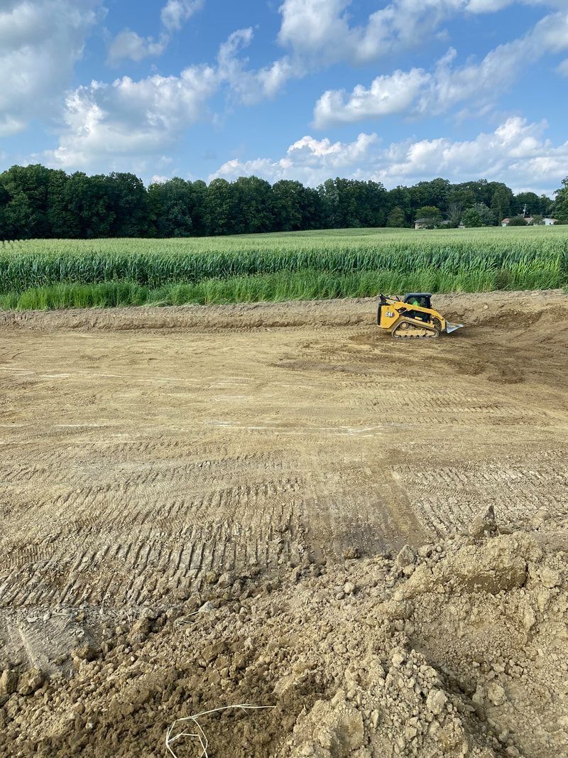 A bulldozer works on a dirt lot, cornfield in the background under a blue sky.