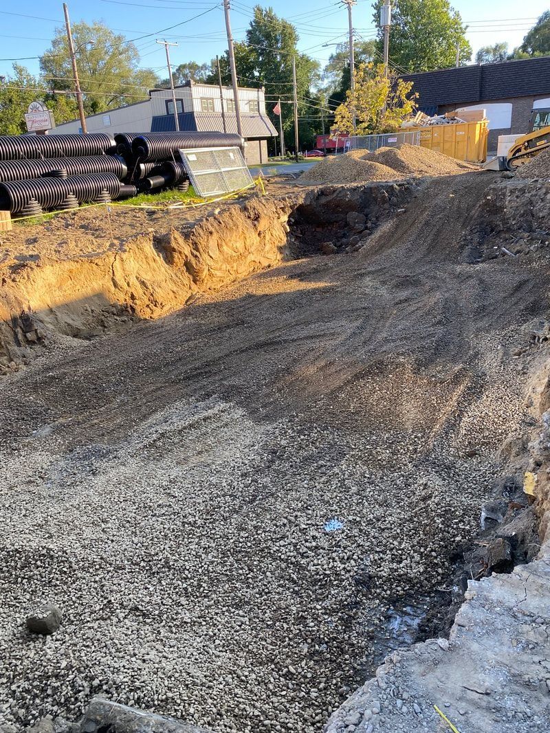Excavated construction site; piles of dirt and gravel; utility poles in background; residential area.