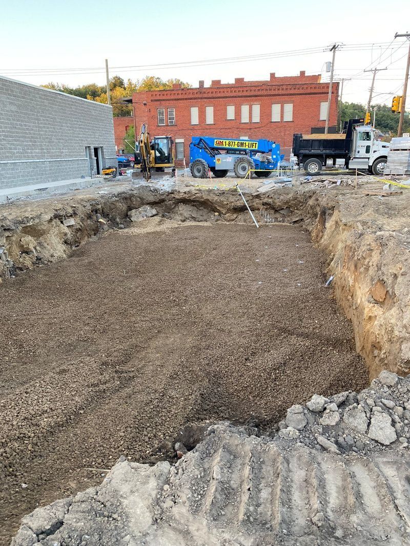 Construction site with excavation and machinery; red brick building in background.
