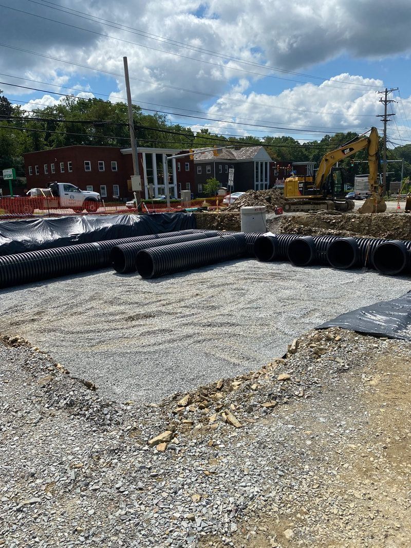 Construction site with gravel, black pipes, and heavy machinery, with buildings and trees in the background under a blue sky.