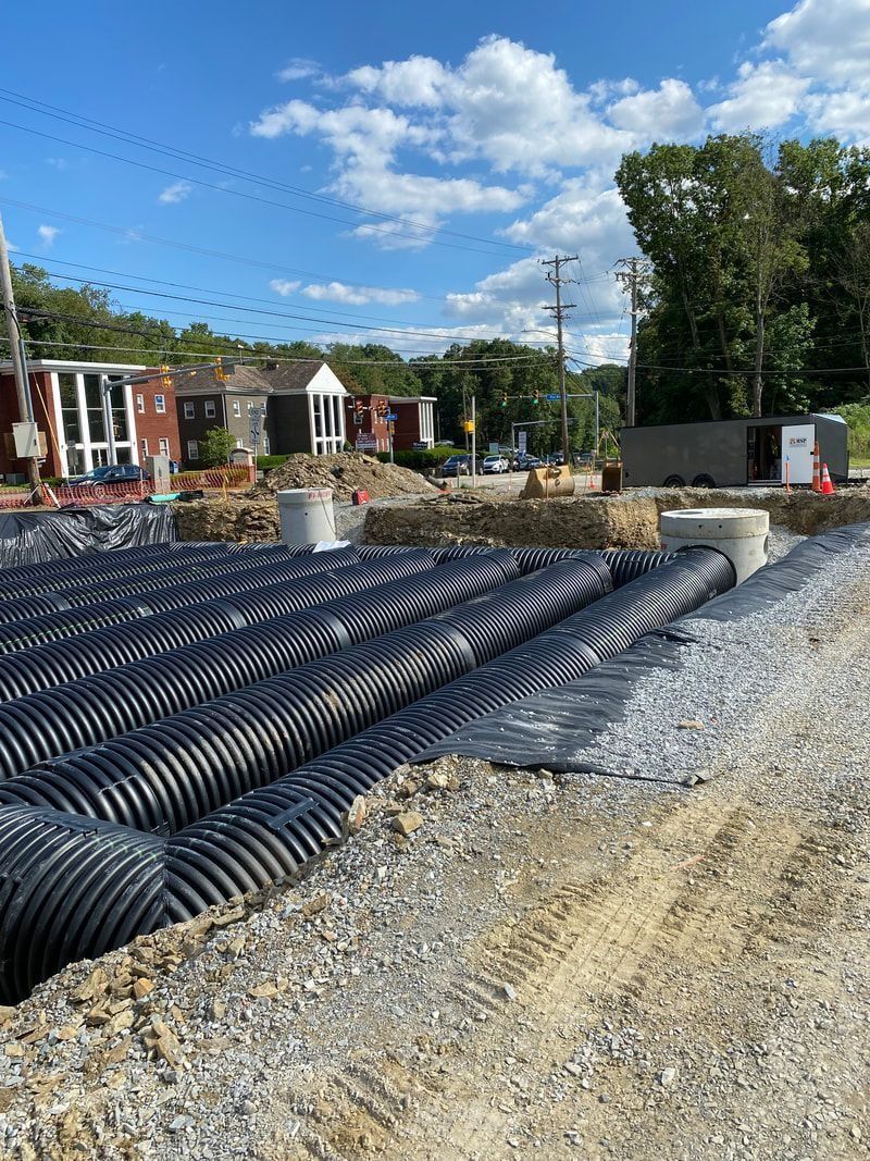 Construction site with corrugated drainage pipes and gravel. Residential buildings in the background under a blue sky.