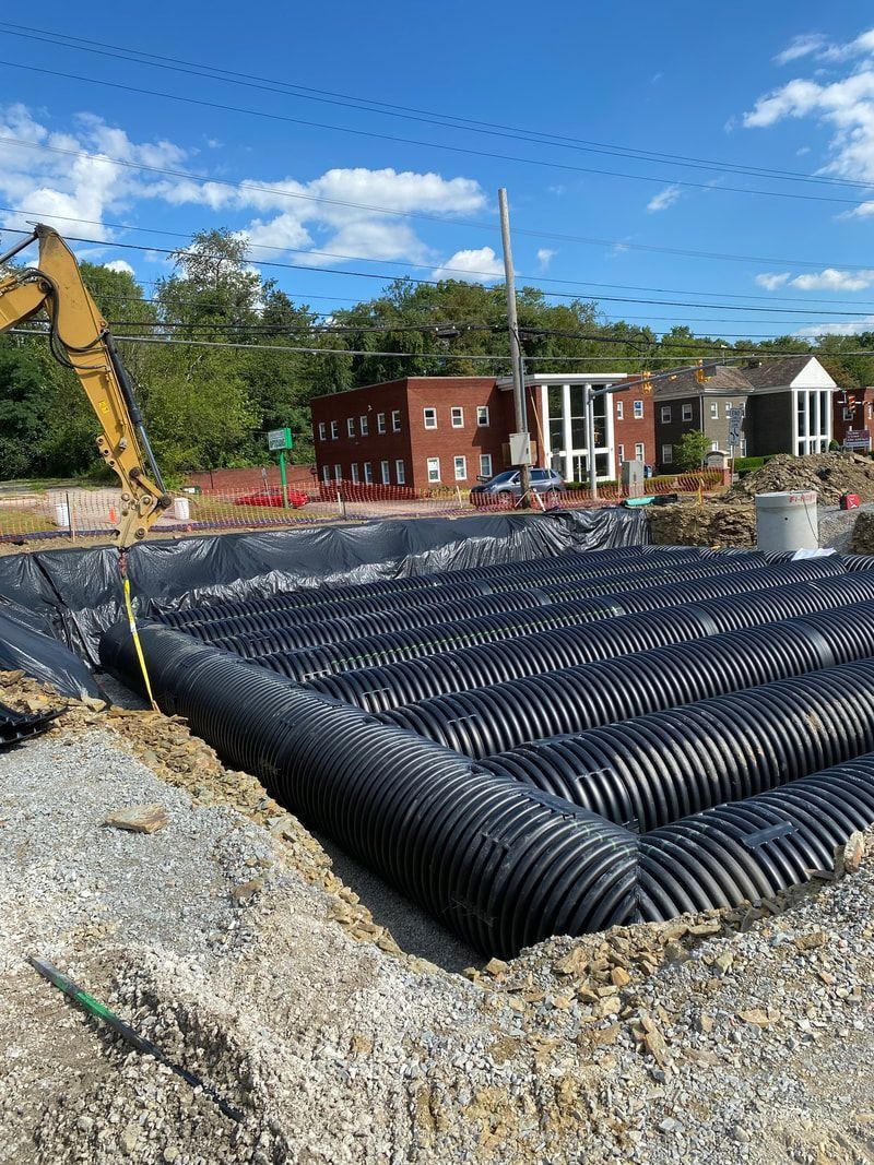 Construction site with black drainage pipes laid out; buildings and excavator in background.