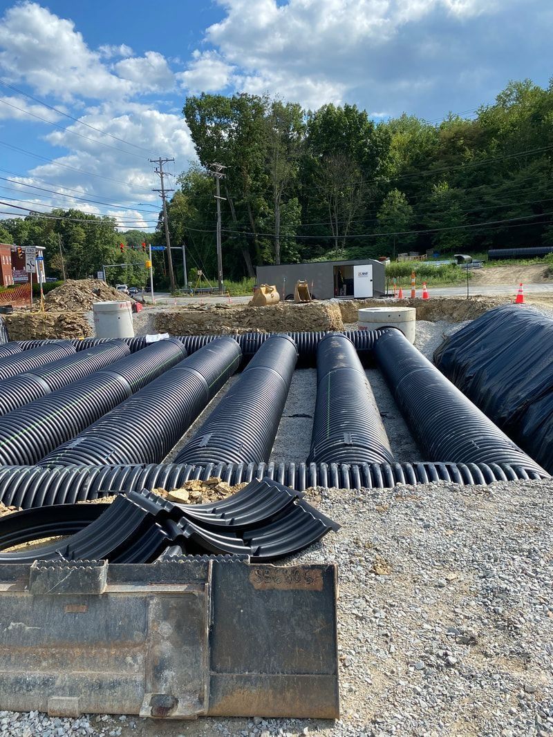 Construction site with large, corrugated black pipes laid in a trench, gravel ground, blue sky with clouds.