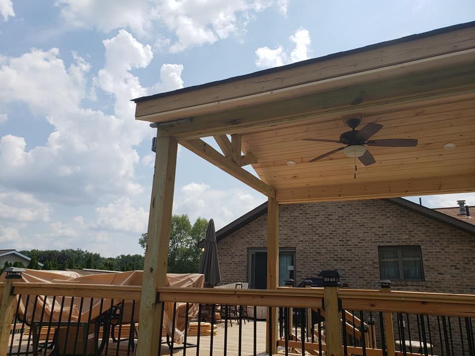 Wooden deck with covered patio, ceiling fan, and black railings, against a brick house and cloudy sky.