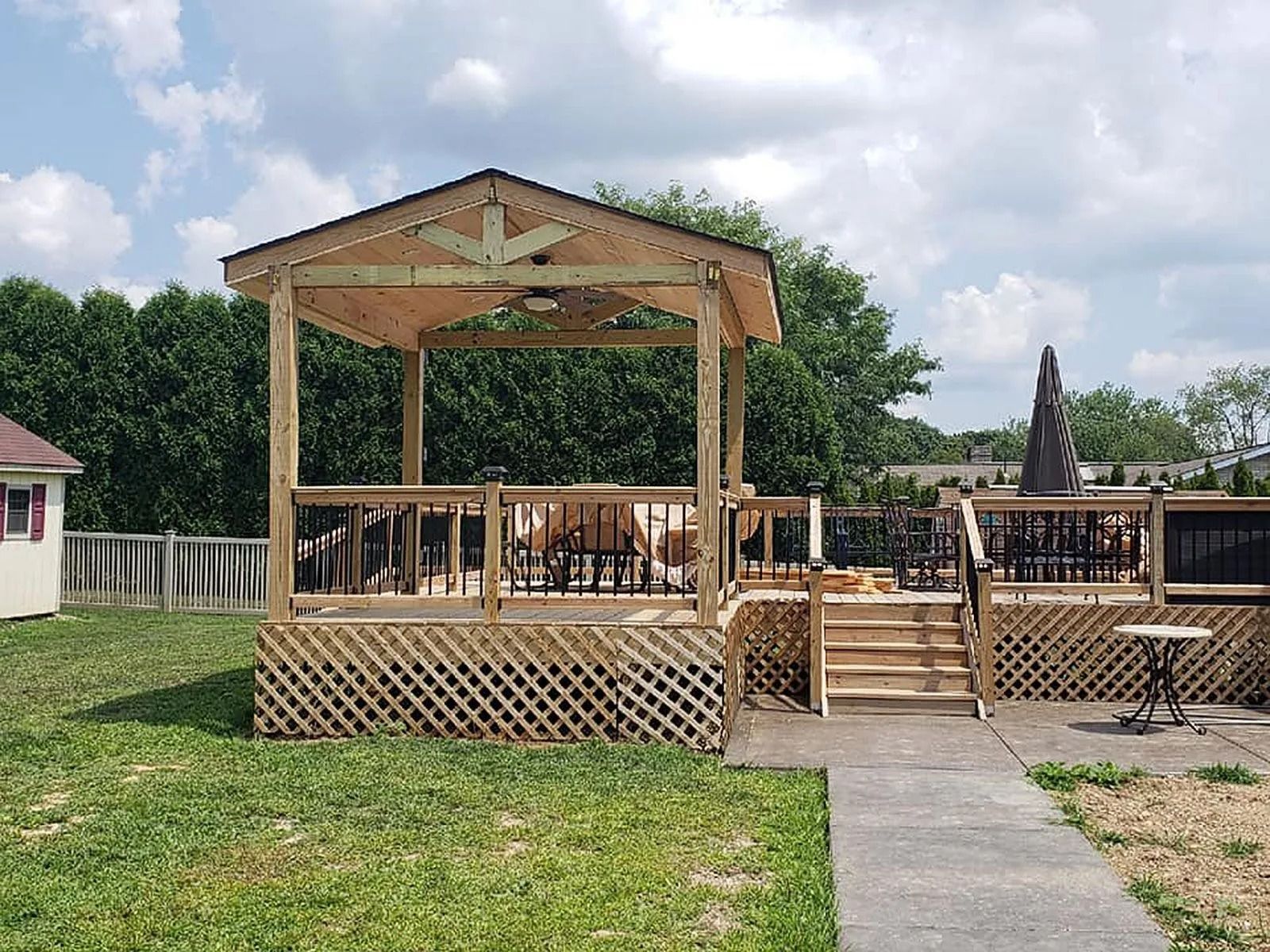 Wooden gazebo on a deck with lattice, leading to an outdoor seating area. Green grass and trees in the background.